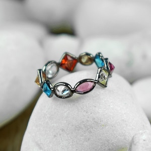 Puede incluir: Un anillo de plata con una banda de piedras preciosas de colores. El anillo está engastado con piedras de diferentes formas ovaladas y cuadradas en tonos de azul, verde, rosa, rojo y amarillo.