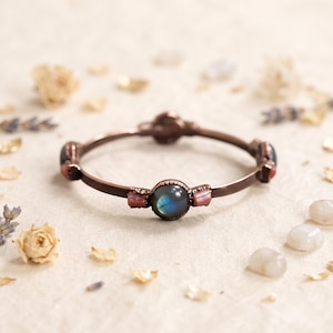 May include: A copper-toned metal bangle bracelet featuring a central round cabochon labradorite gemstone with blue iridescence, flanked by small rectangular pink stones set in decorative metal prongs along the thin circular band.