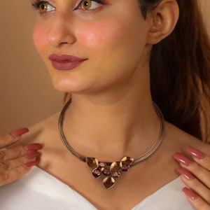 May include: A woman wearing a gold choker necklace with a floral design. The necklace has a delicate chain and a large, intricate pendant with pink accents.