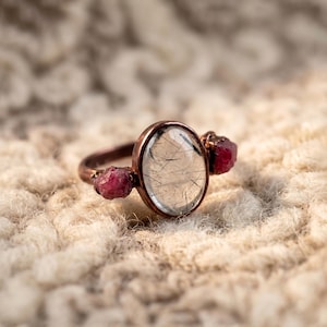 May include: A copper-toned ring featuring an oval, clear stone with black inclusions at the center. Two small, rough-cut, red stones flank the central stone. The ring is set against a textured, off-white background.