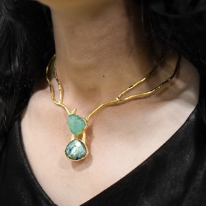 以下が含まれることがあります： 繊細な枝のようなデザインのゴールドネックレス。ネックレスには、緑と青の2つの宝石のペンダントが付いています。