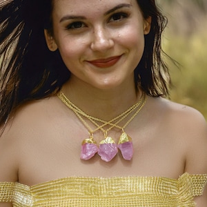 May include: A woman wearing a gold necklace with three pink crystal pendants. The necklace is layered and the crystals are faceted.