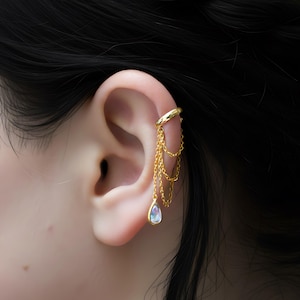 Könnte beinhalten: Ein goldener Ohrklemmen-Schmuck mit Kette und Tropfenanhänger. Der Ohrklemmen-Schmuck umschließt den oberen Teil des Ohrs und ist mit mehreren feinen Ketten verbunden, an denen ein birnenförmiger, schillernder Edelstein hängt.