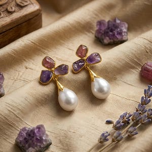 May include: A pair of gold-colored earrings with amethyst and pearl accents. The earrings feature a floral design with purple and pink gemstones and a large white pearl drop. The earrings are displayed on a beige fabric with other gemstones and lavender sprigs.