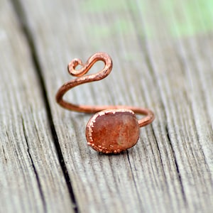 May include: A copper-colored adjustable ring with a reddish-brown stone. The ring features a decorative swirl design and a bezel setting around the stone. The ring is set against a weathered wooden surface.