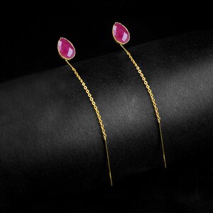 Puede incluir: Un par de pendientes colgantes dorados. Cada pendiente presenta una gema en forma de lágrima, de color rubí, en la parte superior, conectada a una delicada cadena que se extiende hacia abajo. El fondo es negro liso.