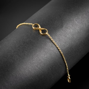 Op de afbeelding: Een gouden armband met een oneindigheidssymbool bedel en een delicate ketting. De armband wordt getoond op een zwarte achtergrond.