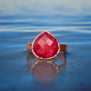 May include: A copper ring with a large, faceted, pink gemstone set in the center. The ring is sitting on a blue surface.