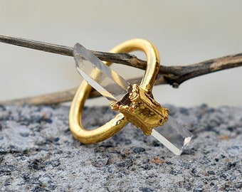 Könnte beinhalten: Ein goldener Ring mit einem klaren Quarzkristall, der in eine goldene Fassung gefasst ist. Der Ring liegt auf einer grauen Oberfläche.