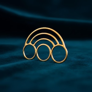 Puede incluir: Broche dorado con forma de arcoíris. El broche presenta tres bandas arqueadas y tres aberturas circulares. La joya se exhibe sobre una superficie de terciopelo verde azulado oscuro, creando un contraste con el oro brillante.