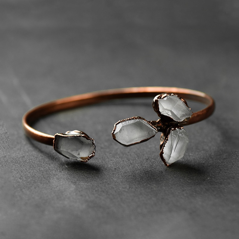 Op de afbeelding: Een koperen armband met vier bevestigde heldere kwartskristallen. De armband is open en heeft een eenvoudig ontwerp.