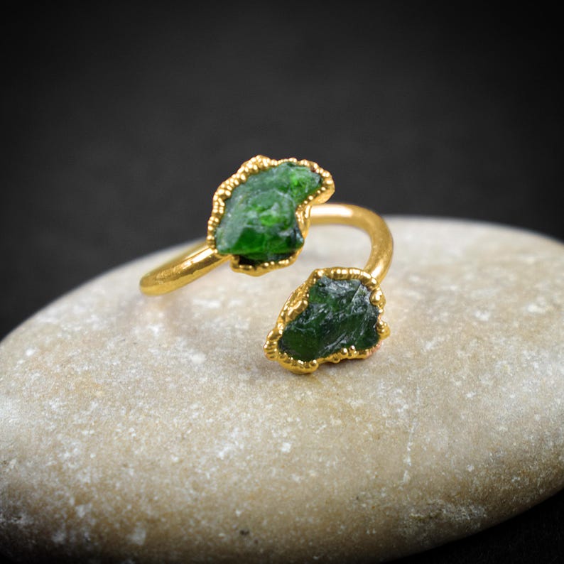 May include: A gold ring with two green gemstone settings. The ring is open and adjustable.