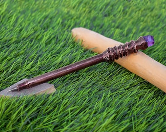 May include: A brown metal pen with a purple gemstone on the top. The pen is lying on a piece of wood and green grass.
