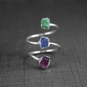 May include: A silver ring featuring three raw gemstones: a green emerald, a blue sapphire, and a purple amethyst. The ring has a spiral band.