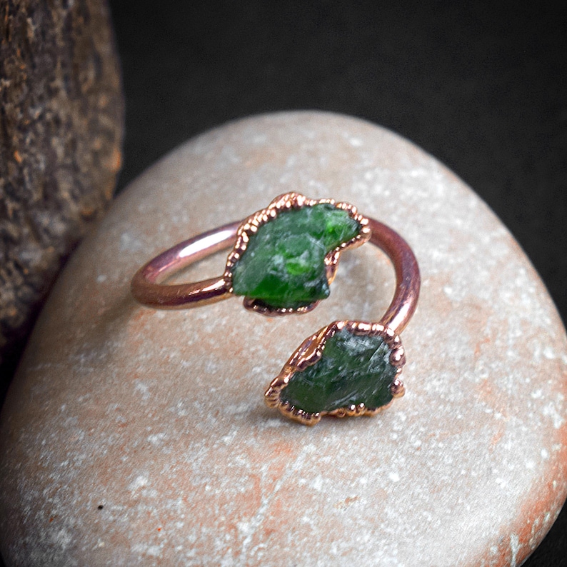 May include: A copper ring with two green gemstone pieces set in the band. The ring is on a light brown stone surface.