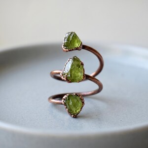 Könnte beinhalten: Ein spiralförmiger Kupferring mit drei rohen, durchscheinenden grünen Peridotsteinen, die in Abständen entlang des Metallbandes angebracht sind. Das Kupfer hat ein warmes, rotbraunes Metallic-Finish und die Steine haben unebene Kanten.