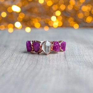 May include: A rose gold ring set with five faceted gemstones. The centre stone is clear and the four surrounding stones are a deep pink.