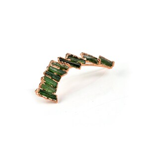 Puede incluir: Un pendiente de aro de oro con un dise&ntilde;o curvo que presenta una fila de piedras preciosas verdes.