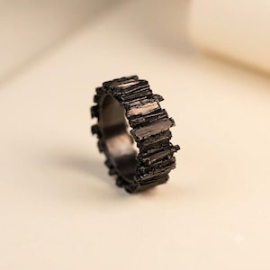 Könnte beinhalten: Ein schwarzer Ring mit einer texturierten, gezackten Außenfläche, die aus vertikalen, splitterartigen Segmenten besteht. Das Band hat ein dunkles, mattes Finish mit unregelmäßigen Kanten, die ein raues, unebenes Profil um den Ring bilden.