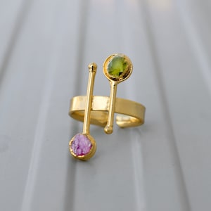 May include: Gold ring with two stones, one green and one pink. The ring is designed with two thin gold bars that extend from the band and hold the stones.