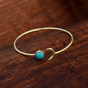 Puede incluir: Un brazalete de oro con una piedra turquesa y un dije de media luna.