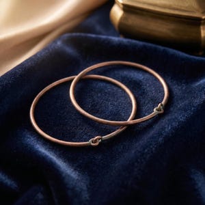 May include: Two circular copper-toned bangles with a smooth, thin metal band construction. Each bangle features a small, integrated loop closure where the ends of the wire meet, creating a simple, continuous circular form with a metallic finish.