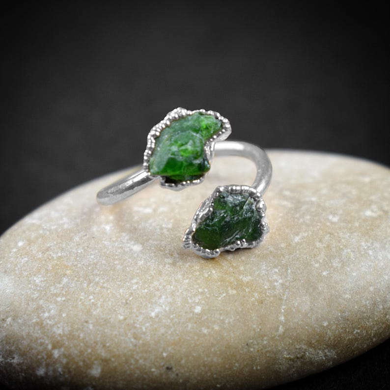 May include: A silver ring with two green gemstone settings. The ring is designed to wrap around the finger.