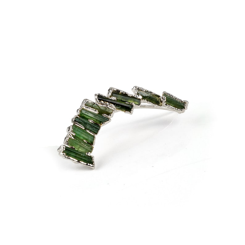 Puede incluir: Un pendiente de plata con un dise&ntilde;o curvo adornado con m&uacute;ltiples piedras preciosas verdes.