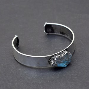 May include: A silver cuff bracelet featuring a large, rough-cut blue gemstone set into the metal. The bracelet is open and ready to be worn.