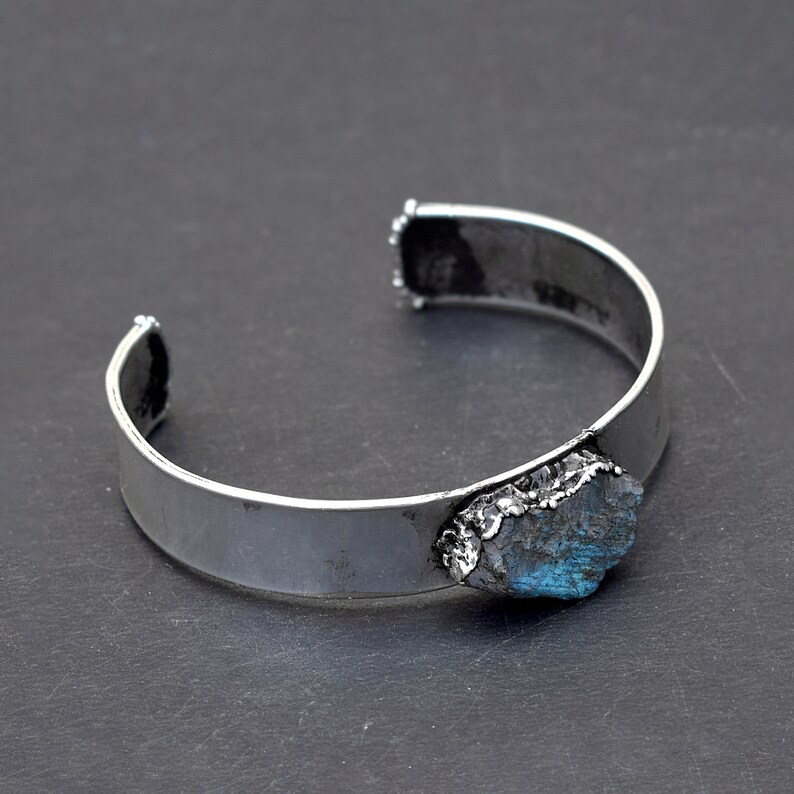 May include: A silver cuff bracelet featuring a large, rough-cut blue gemstone set into the metal. The bracelet is open and ready to be worn.
