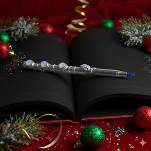 Op de afbeelding: Een zilveren pen met een decoratief ontwerp en opalescente stenen ligt op een open zwart notitieboek met roodgelijnde pagina's. De pen is omgeven door kerstversieringen en feestelijke decoraties op een rode fluwelen ondergrond.