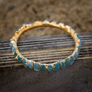 May include: A gold bangle bracelet with a row of light blue, faceted gemstones. The bracelet is on a wooden surface.