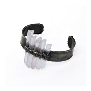 May include: A silver cuff bracelet featuring a cluster of clear quartz crystals set into the metal. The crystals are arranged in a staggered pattern, creating a unique and eye-catching design.