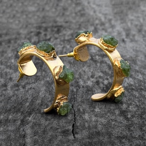 Puede incluir: Aretes de aro dorados con detalles de piedras preciosas verdes. Los aretes están hechos de metal y tienen un acabado pulido.