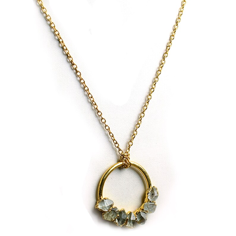 Puede incluir: Un collar de cadena dorada con un colgante circular que presenta un grupo de peque&ntilde;as piedras preciosas grises sin pulir.