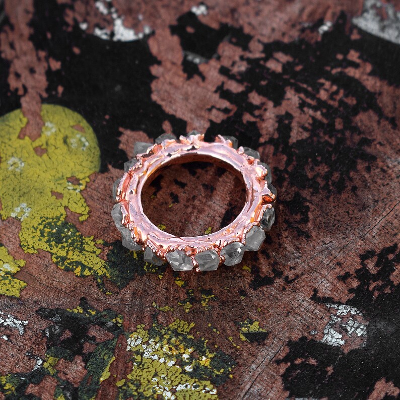 Puede incluir: Un anillo de oro rosa con una banda de cristales de cuarzo transparentes. El anillo est&aacute; colocado sobre una superficie marr&oacute;n texturizada.