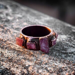 Pode incluir: Um anel de cobre com uma banda adornada com v&aacute;rias pedras preciosas rosa e roxas. O anel est&aacute; sobre uma superf&iacute;cie &aacute;spera e cinza.