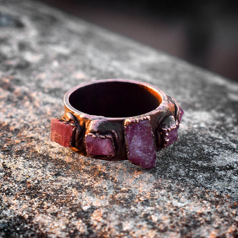 Pode incluir: Um anel de cobre com uma banda adornada com v&aacute;rias pedras preciosas rosa e roxas. O anel est&aacute; sobre uma superf&iacute;cie &aacute;spera e cinza.