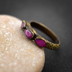 May include: A gold ring with three pink gemstones set in the band. The band is decorated with a floral design.