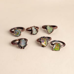 May include: Six rings featuring raw, iridescent opal gemstones set in dark, textured copper bands. Each ring showcases a unique, irregular stone with flashes of green, orange, and blue color, held by a rugged, organic metal setting with a dark finish.