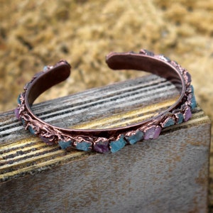 May include: A copper bracelet with a row of blue and pink gemstones. The bracelet is on a wooden surface.