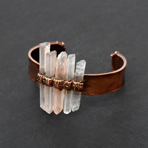 May include: A copper cuff bracelet with clear quartz crystals attached. The crystals are arranged in a staggered pattern and are held in place by copper wire.