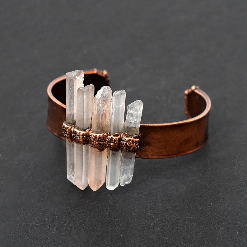 May include: A copper cuff bracelet with clear quartz crystals attached. The crystals are arranged in a staggered pattern and are held in place by copper wire.
