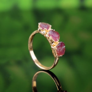 May include: A gold ring with three red gemstone settings. The gemstones are faceted and have a rough, natural look.