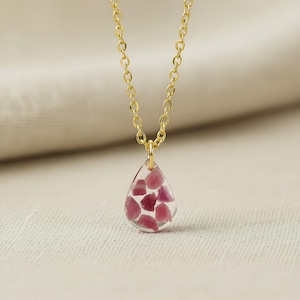 May include: A gold-colored chain necklace with a teardrop pendant. The pendant is clear with embedded pink-red stones. The necklace is displayed on a neutral-colored fabric background. Jewelry.