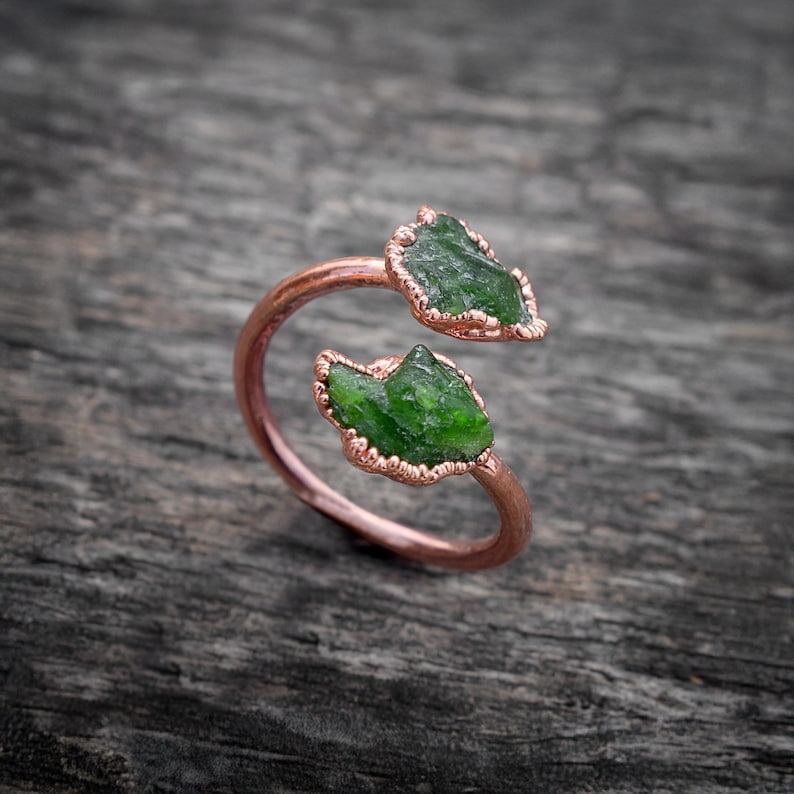 May include: A rose gold ring with two green gemstone accents. The ring is open and adjustable.