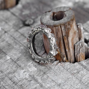Puede incluir: Un anillo de plata con un dise&ntilde;o &uacute;nico que presenta varias piedras peque&ntilde;as y transparentes. El anillo est&aacute; apoyado en un trozo de madera desgastada.