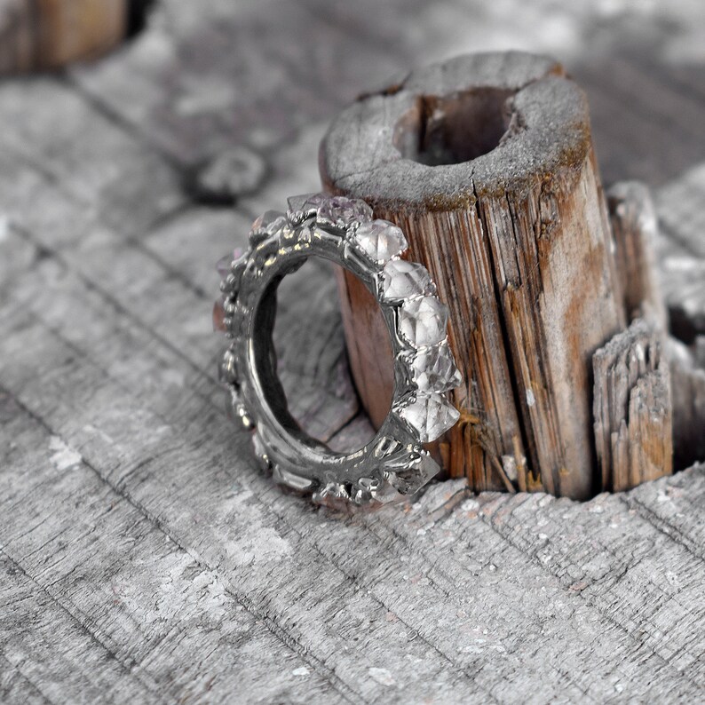 Puede incluir: Un anillo de plata con un dise&ntilde;o &uacute;nico que presenta varias piedras peque&ntilde;as y transparentes. El anillo est&aacute; apoyado en un trozo de madera desgastada.