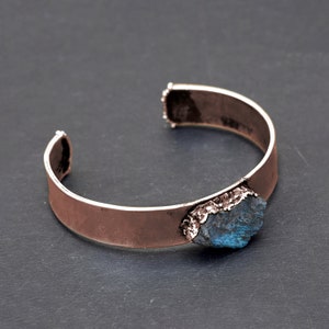 May include: A copper cuff bracelet featuring a large, blue-green gemstone set into the metal. The gemstone has a rough, natural texture.