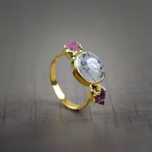 Puede incluir: Un anillo dorado con una piedra grande, redonda y transparente con inclusiones negras, flanqueada por dos piedras rosas más pequeñas y en bruto. La banda y el engaste del anillo son dorados, creando un contraste llamativo con las piedras preciosas.